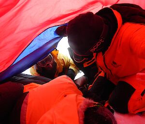 One man lays inside a red tent with two men kneeling next to him