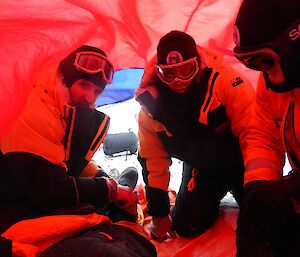 Three men kneel inside a red bivvy bag.