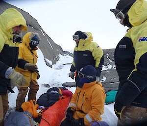 Five men in yellow jackets stand around a patient in a rescue sled