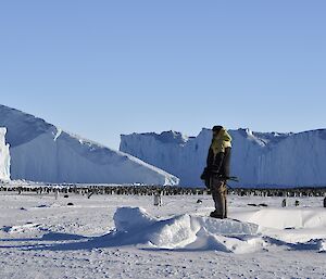 A man stands on ice with emperor penguins and icebergs in the background.