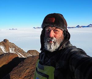 A man has a frozen beard and moustache standing on top of a mountain surrounded by a white plateau