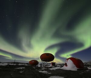 White and green rays appear in the night sky above three red domed huts.