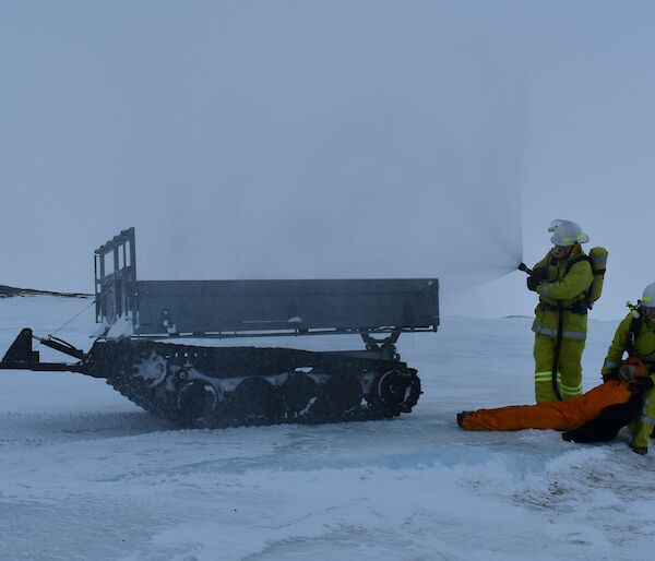 Two men in fire fighting gear retrieve a dummy and use a fire hose on ice.