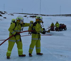 Two men stand on ice in fire fighting gear holding a fire hose.
