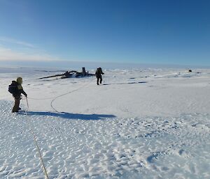 Two people connected by ropes walk across ice towards a wreck on the glacier