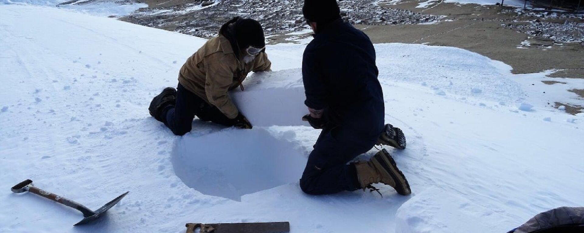 Two men cut ice blocks from an ice embankment.