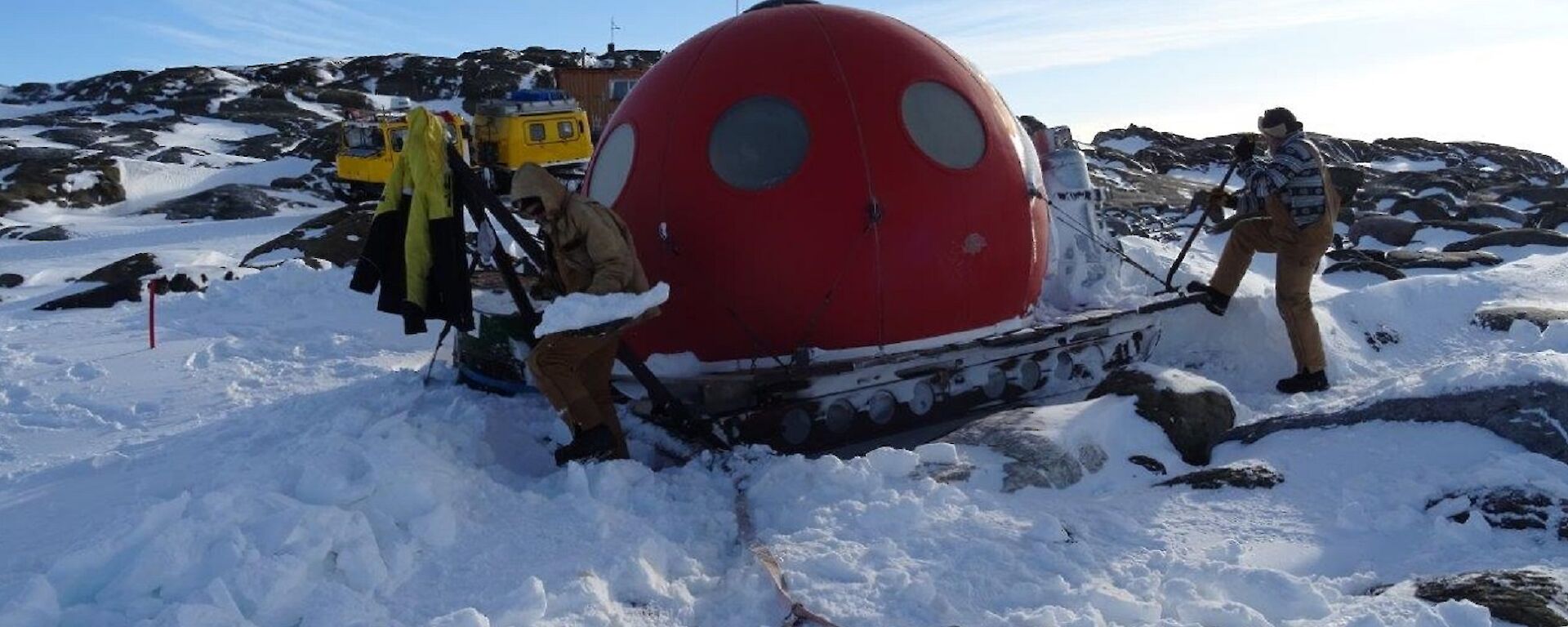 Two men work on a red domed shaped hut in the snow