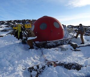 Two men work on a red domed shaped hut in the snow