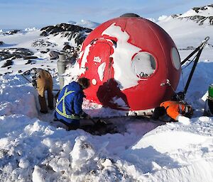Three men are digging around a red domed hut in the snow