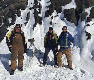 Three men stand in front of a camera on a tripod on an island covered in snow.