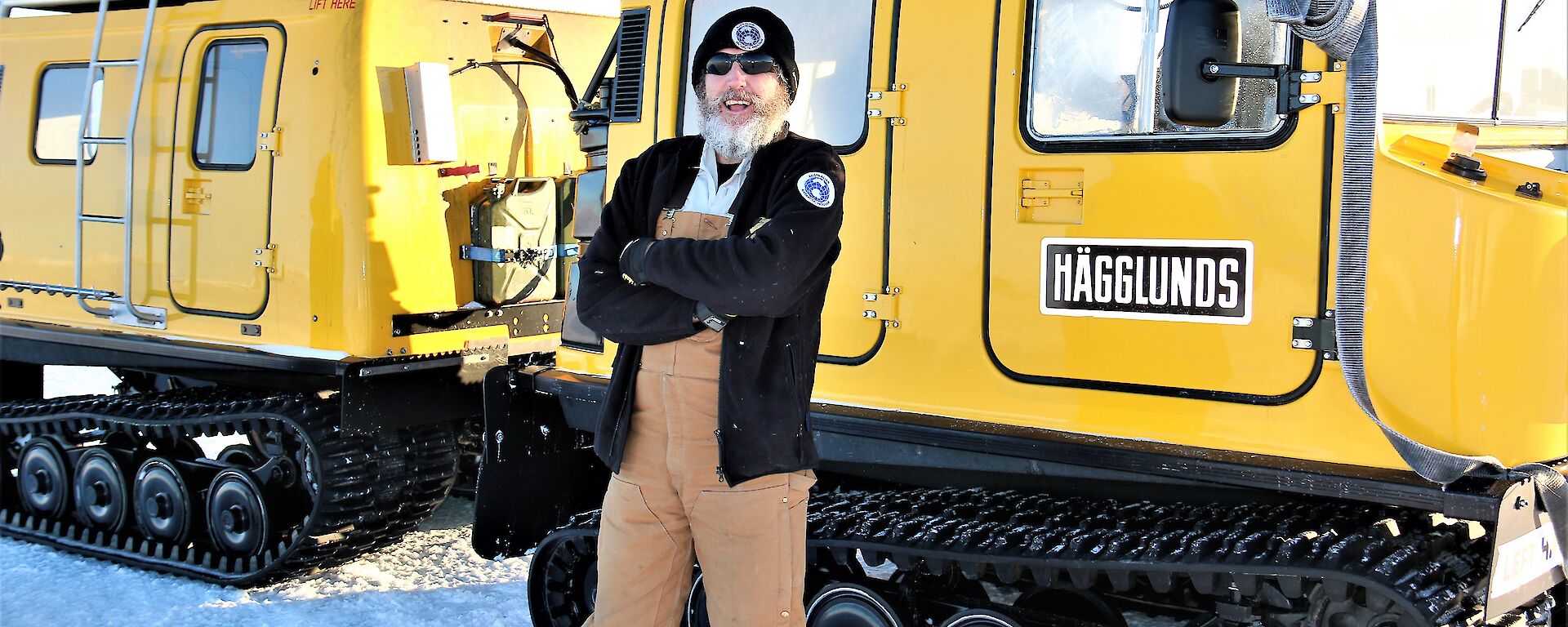 A man wearing sunglasses and a beanie stands in front of a yellow Hägglunds