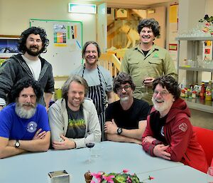 Seven men sit around a table, five wearing wigs.