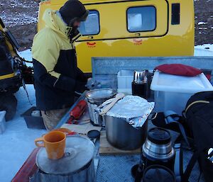 A man holds a coffee percolator in front of an outdoor camp kitchen