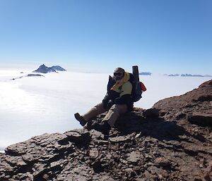 A man sits on the edge of a rocky mountain range with mountain range and Nunatak peaks in the background