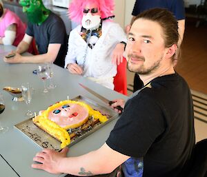 A young man looks at the camera in front of a birthday cake with people in costume in the background