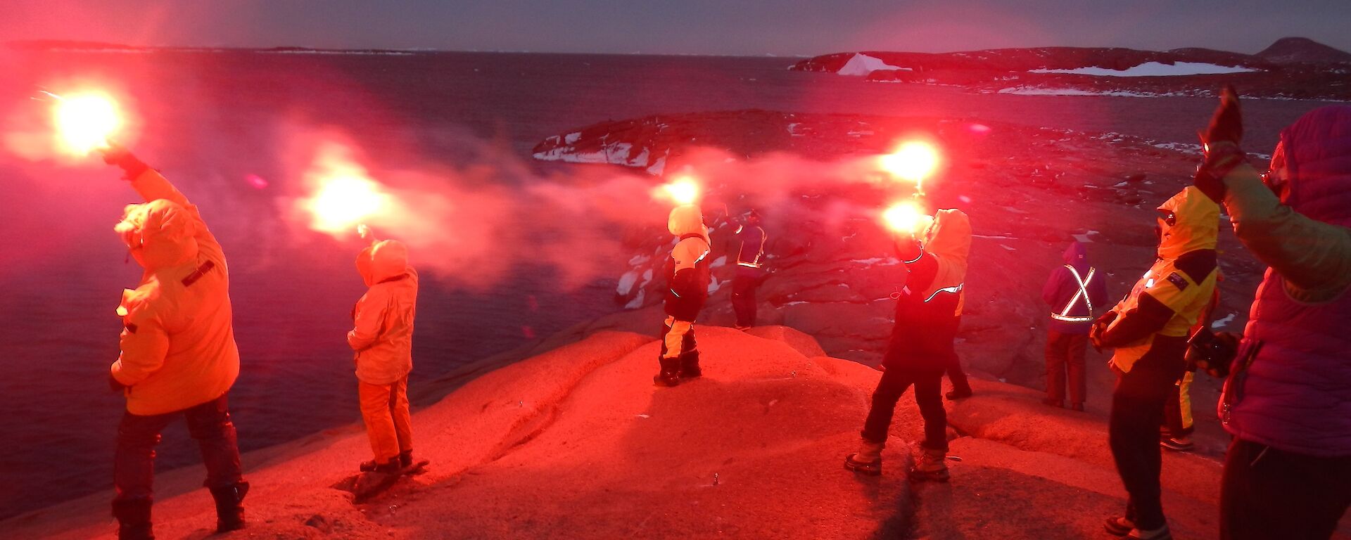 A group of people stand on a rock peninsula holding red flares in the sky.