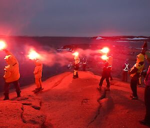 A group of people stand on a rock peninsula holding red flares in the sky.