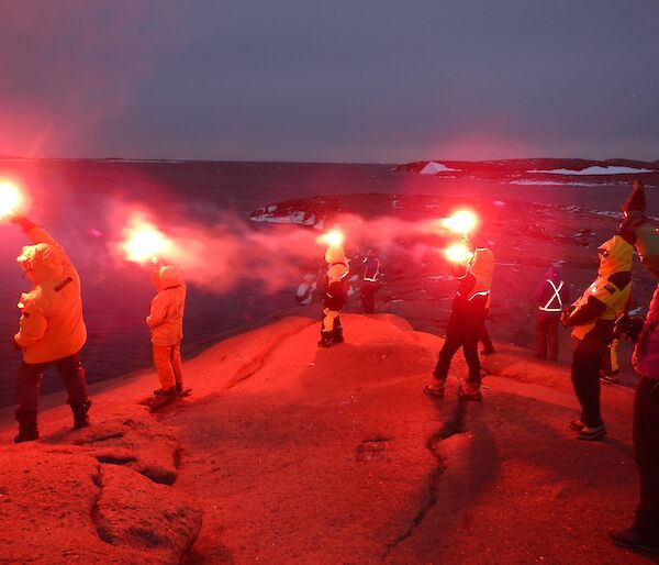 A group of people stand on a rock peninsula holding red flares in the sky.