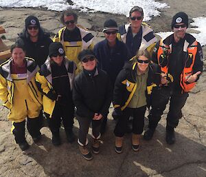 Nine people in Antarctic clothing look up at the camera while standing outside.