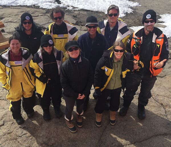 Nine people in Antarctic clothing look up at the camera while standing outside.