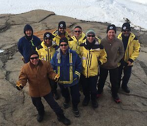 Nine men in Antarctic clothing look up at the camera while standing outside.
