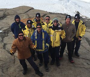 Nine men in Antarctic clothing look up at the camera while standing outside.