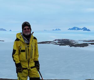 A man in a yellow freezer suit stands on top of an island with sea ice in background
