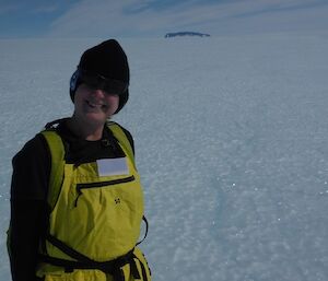 A woman wearing a beanie stands on an icy plateau with a mountain range behind her.