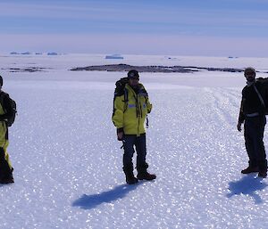 Three men stand on an icy plateau with islands and icebergs behind them.