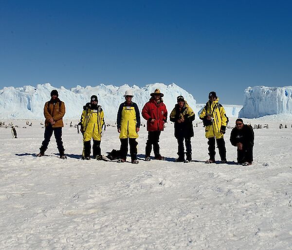 7 people are lined up in front of a penguin rookery with icebergs in the background.