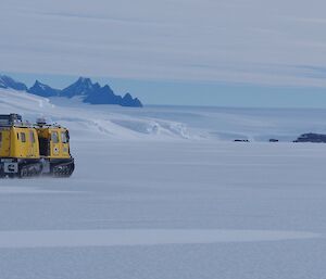 A yellow Hägglunds drives along the sea ice with a mountain range in the background.