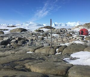 A rocky island with a colony of Adelie penguins, a red melon hut and icebergs in the background.