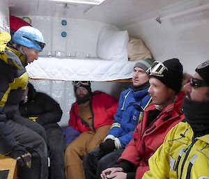Six people are seated inside a small van wearing Antarctic clothing.