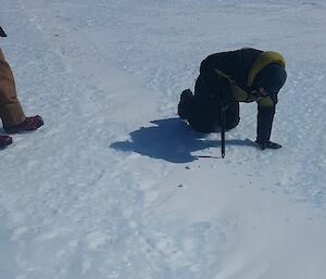 A person is kneeling on the snow pushing an ice axe into the snow.