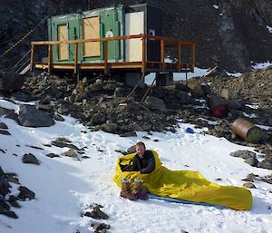 A man lays in a yellow plastic bag in front of a field hut.