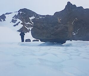 An expeditioner stands next to a moraine boulder frozen on top of a pile of ice.