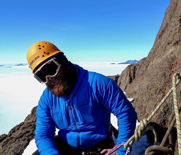 A man wearing a blue jacket sits on the side of a mountain holding ropes.