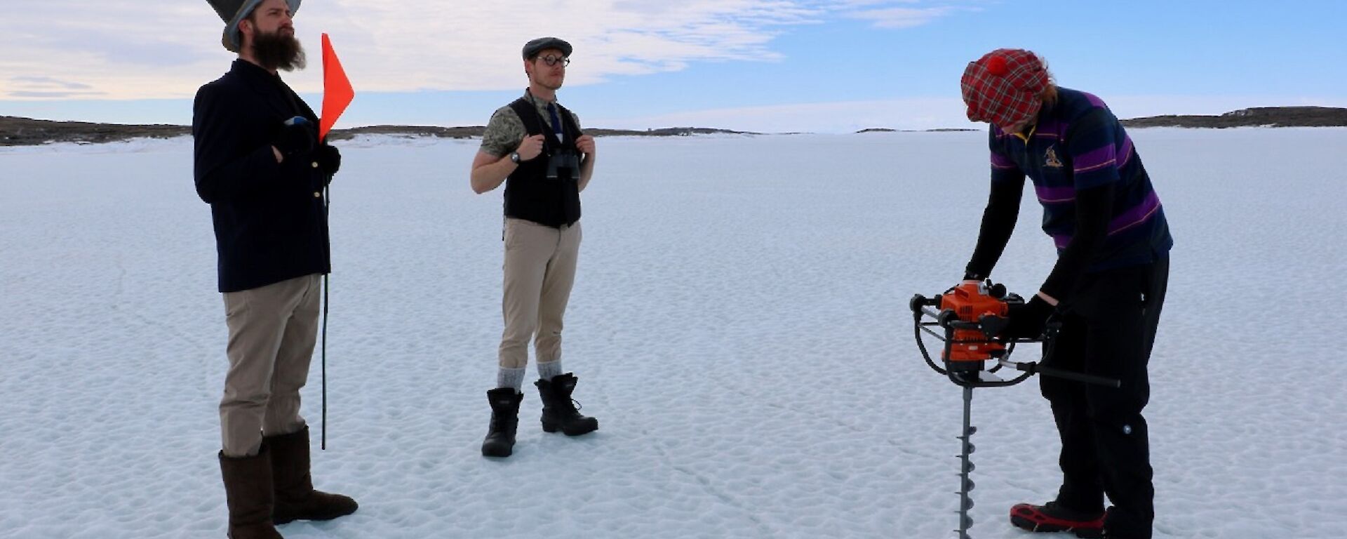 Three men stand on sea ice in golf attire. One man drills a hold in the sea ice