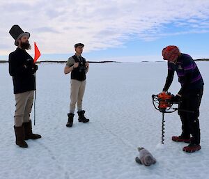 Three men stand on sea ice in golf attire. One man drills a hold in the sea ice