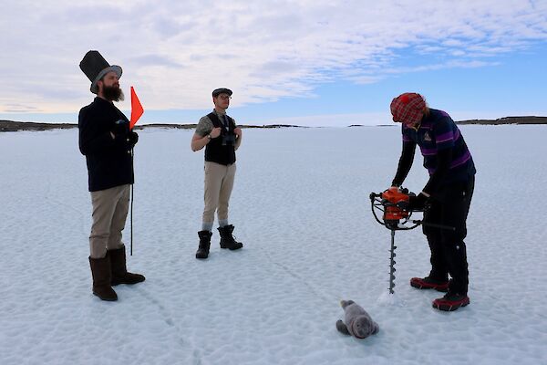 Three men stand on sea ice in golf attire. One man drills a hold in the sea ice