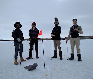 Four men stand on sea ice in golf attire with golf clubs and flag