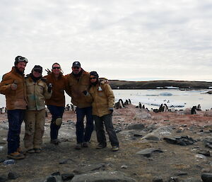 5 people on a beach smiling at camera with penguin chicks in the background