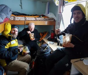 3 men in a field hut eating boil-in-a-bag dinner