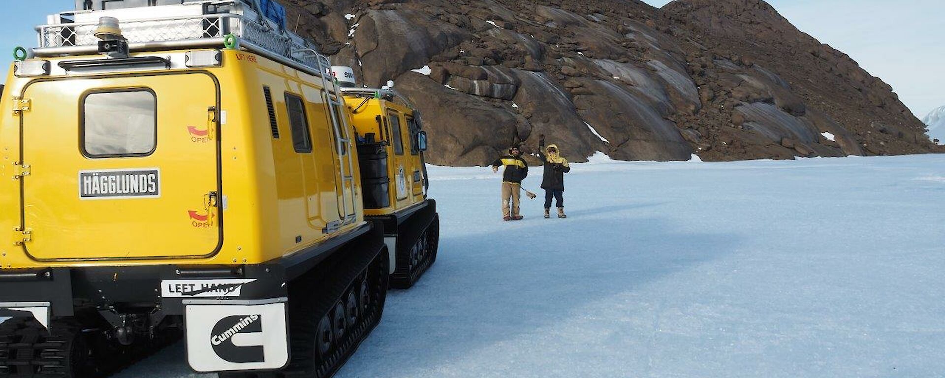 2 men stand on the ice plateau with a Hägglunds vehicle