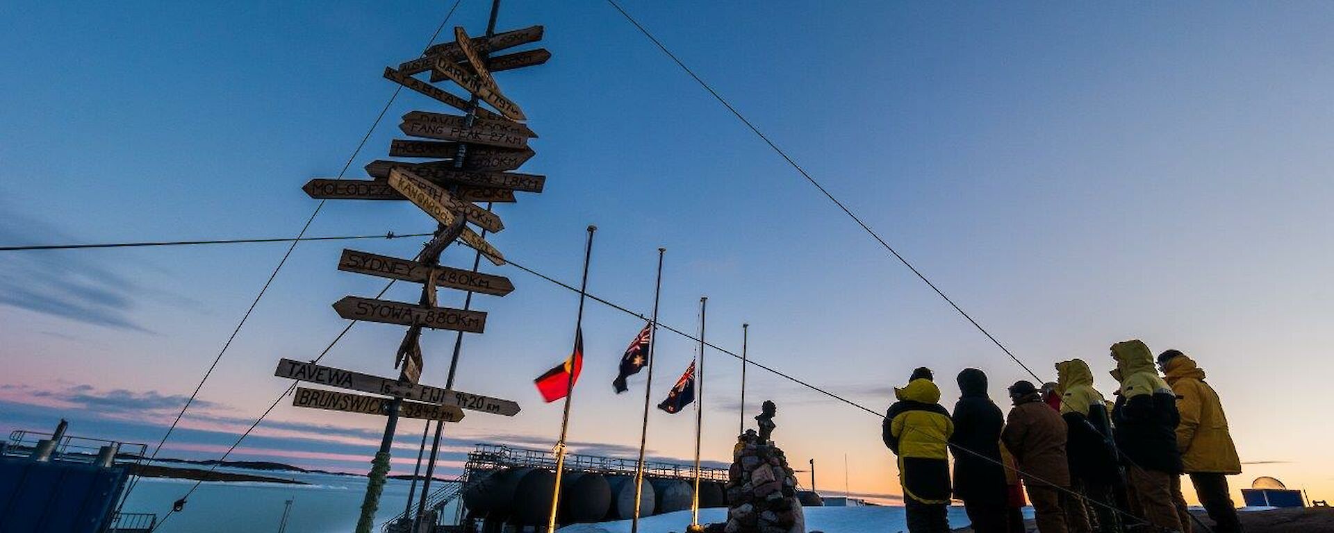 A group of people around flag poles and Mawson station at dawn