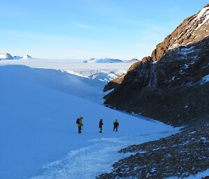 3 people walk down a snowy slope next to a wind scour