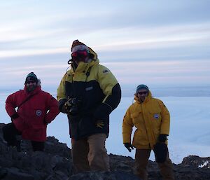 3 men pose on a rocky hill, ice plateau behind.