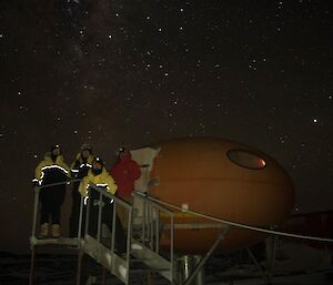 A goupr of people stand outside the hut on a starry night