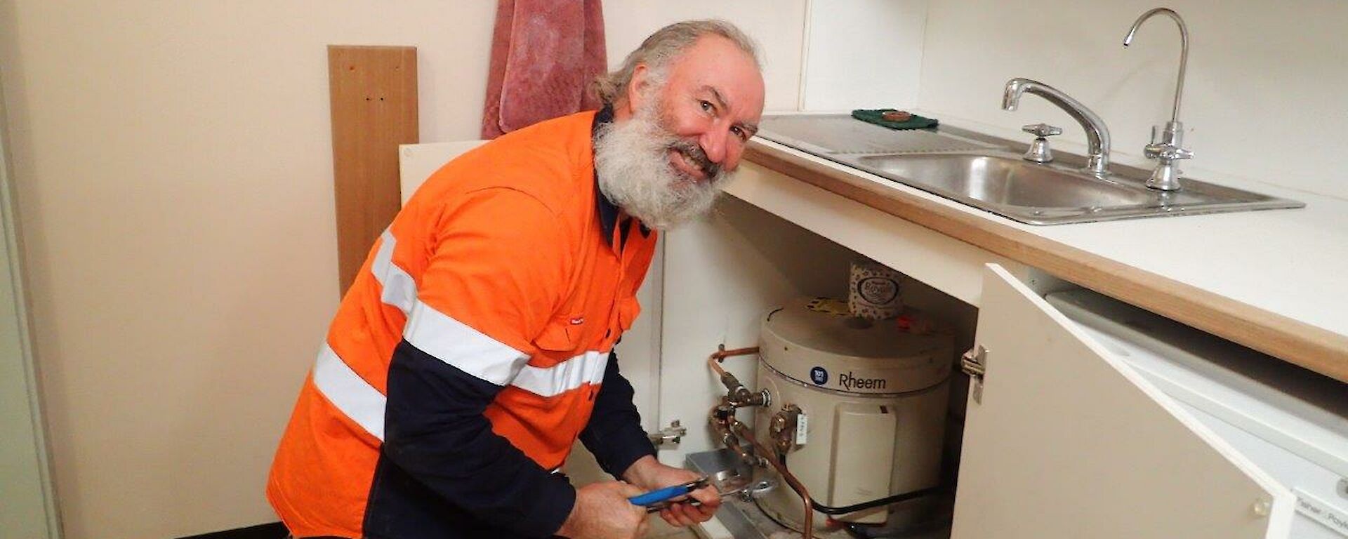 A man smiles at camera as he works on under sink plumbing