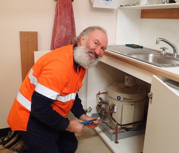 A man smiles at camera as he works on under sink plumbing